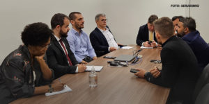 As imagens mostram uma reunião com um grupo de oito pessoas sentadas ao redor de uma mesa de madeira em uma sala de conferências. Elas estão discutindo e tomando notas, todos com uma aparência formal, usando trajes sociais. O ambiente é simples, com uma mesa retangular, cadeiras e paredes claras. Em uma das imagens, há um texto sobreposto que diz: "Após atuação intensa dos Sindicatos, TJMG anuncia avanços importantes para a categoria".