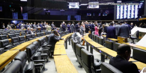 Vista lateral do Plenário da Câmara dos Deputados, há diversas fileiras de cadeiras pretas vazias, ao fundo há um grupo com muitas pessoas vestidas com trajes sociais, à frente deles está a mesa diretiva da Câmara. Conteúdo textual: NÃO AO RRF-Relator apresenta parecer favorável ao Propag e proposta avança na Câmara dos Deputados.