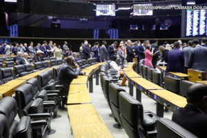 Vista lateral do Plenário da Câmara dos Deputados, há diversas fileiras de cadeiras pretas vazias, ao fundo há um grupo com muitas pessoas vestidas com trajes sociais, à frente deles está a mesa diretiva da Câmara. Conteúdo textual: NÃO AO RRF-Relator apresenta parecer favorável ao Propag e proposta avança na Câmara dos Deputados.