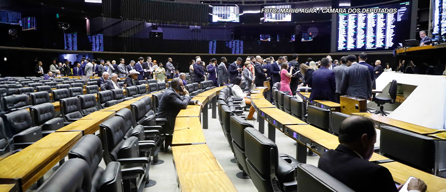 Vista lateral do Plenário da Câmara dos Deputados, há diversas fileiras de cadeiras pretas vazias, ao fundo há um grupo com muitas pessoas vestidas com trajes sociais, à frente deles está a mesa diretiva da Câmara. Conteúdo textual: NÃO AO RRF-Relator apresenta parecer favorável ao Propag e proposta avança na Câmara dos Deputados.