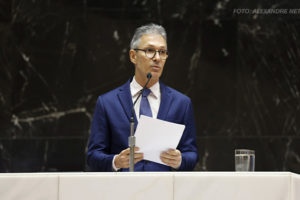 A imagem mostra o governador de Minas Gerais, Romeu Zema, vestido com um terno azul e gravata, falando em um púlpito enquanto segura um papel. Ele está diante de um fundo escuro de mármore e há um microfone à sua frente. O título em destaque menciona que ele se comprometeu a aderir ao programa Propag, enquanto sindicatos cobram rapidez no processo.