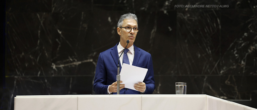 A imagem mostra o governador de Minas Gerais, Romeu Zema, vestido com um terno azul e gravata, falando em um púlpito enquanto segura um papel. Ele está diante de um fundo escuro de mármore e há um microfone à sua frente. O título em destaque menciona que ele se comprometeu a aderir ao programa Propag, enquanto sindicatos cobram rapidez no processo.