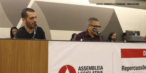 A imagem mostra uma cena de debate público realizado na Assembleia Legislativa de Minas Gerais, onde o coordenador-geral do SINJUS, Alexandre Pires, participa de uma discussão contra o desmonte do Ipsemg. Na parte superior há pessoas sentadas, com microfones, em um ambiente de debate formal