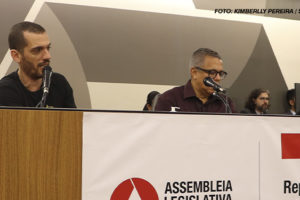 A imagem mostra uma cena de debate público realizado na Assembleia Legislativa de Minas Gerais, onde o coordenador-geral do SINJUS, Alexandre Pires, participa de uma discussão contra o desmonte do Ipsemg. Na parte superior há pessoas sentadas, com microfones, em um ambiente de debate formal