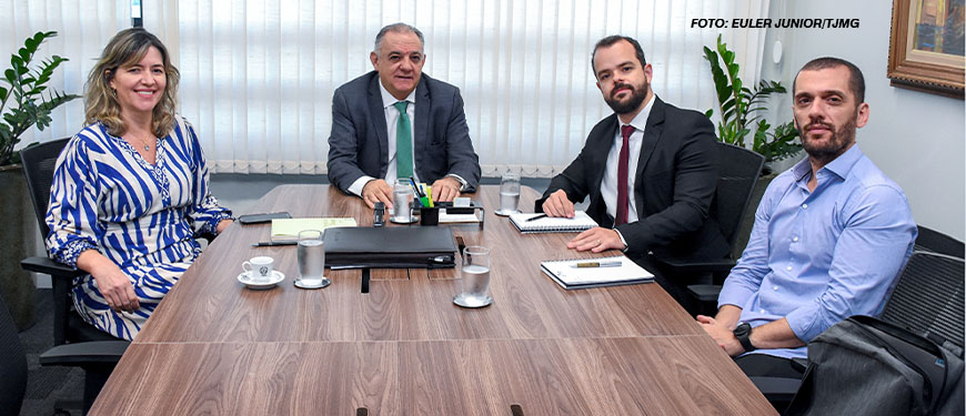 A imagem mostra uma reunião em uma sala de conferências, onde há dois representantes da administração do TJMG e dois diretores do SINJUS sentados ao redor de uma mesa de madeira. Ao fundo, é possível ver janelas com persianas fechadas e algumas plantas ornamentais. Estão presentes à mesa o 1º vice-presidente do TJMG, desembargador Marcos Lincoln, Alexandre Pires (coordenador-geral do SINJUS-MG) e Felipe Rodrigues (diretor de Assuntos Jurídicos).