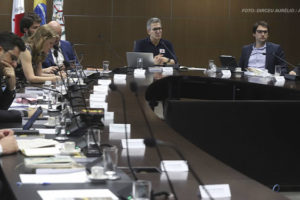 Uma reunião está acontecendo em uma sala de conferências com uma grande mesa em formato de "U". Diversas pessoas, homens e mulheres, estão sentadas ao redor da mesa, olhando para documentos, laptops ou prestando atenção à conversa. Algumas delas estão fazendo anotações. Ao centro da mesa, está Romeu Zema, o governador de Minas Gerais, ele é grisalho, usa óculos e veste uma camisa de manga longa azul marinho.