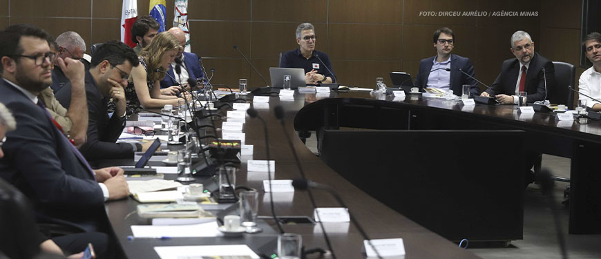 Uma reunião está acontecendo em uma sala de conferências com uma grande mesa em formato de "U". Diversas pessoas, homens e mulheres, estão sentadas ao redor da mesa, olhando para documentos, laptops ou prestando atenção à conversa. Algumas delas estão fazendo anotações. Ao centro da mesa, está Romeu Zema, o governador de Minas Gerais, ele é grisalho, usa óculos e veste uma camisa de manga longa azul marinho.