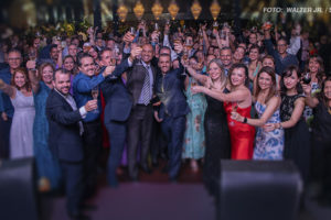 As imagens mostram uma confraternização, com diversas pessoas em trajes formais reunidas e celebrando em um salão iluminado e elegante. Os participantes estão sorrindo e levantando taças em um brinde coletivo, transmitindo um clima de alegria e união. A ocasião é descrita como uma festa de fim de ano organizada pelo SINJUS, celebrando a integração entre os filiados. O ambiente é sofisticado, com decoração requintada, incluindo lustres brilhantes ao fundo. O destaque visual está no grupo de pessoas na frente, exibindo expressões animadas e gestos de confraternização.