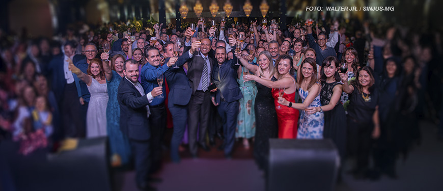 As imagens mostram uma confraternização, com diversas pessoas em trajes formais reunidas e celebrando em um salão iluminado e elegante. Os participantes estão sorrindo e levantando taças em um brinde coletivo, transmitindo um clima de alegria e união. A ocasião é descrita como uma festa de fim de ano organizada pelo SINJUS, celebrando a integração entre os filiados. O ambiente é sofisticado, com decoração requintada, incluindo lustres brilhantes ao fundo. O destaque visual está no grupo de pessoas na frente, exibindo expressões animadas e gestos de confraternização.