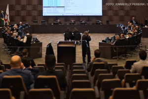 A imagem retrata um auditório formal, amplo e moderno, configurado para uma sessão oficial. Ao fundo, há um painel eletrônico com informações projetadas, acompanhado de uma bancada elevada onde estão reunidos membros do Órgão Especial. Nas laterais e no centro da sala, pessoas estão sentadas em mesas ou cadeiras, em atividades como anotações ou observação, enquanto outros estão em pé registrando o evento. Algumas bandeiras do Brasil e de estados estão dispostas à esquerda, reforçando o caráter institucional. A iluminação é clara, e o espaço está bem organizado, com cadeiras ocupadas por espectadores. A cena transmite o ambiente de uma deliberação oficial importante, destacando a aprovação de mudanças na lei do Auxílio-Saúde para a ALMG.