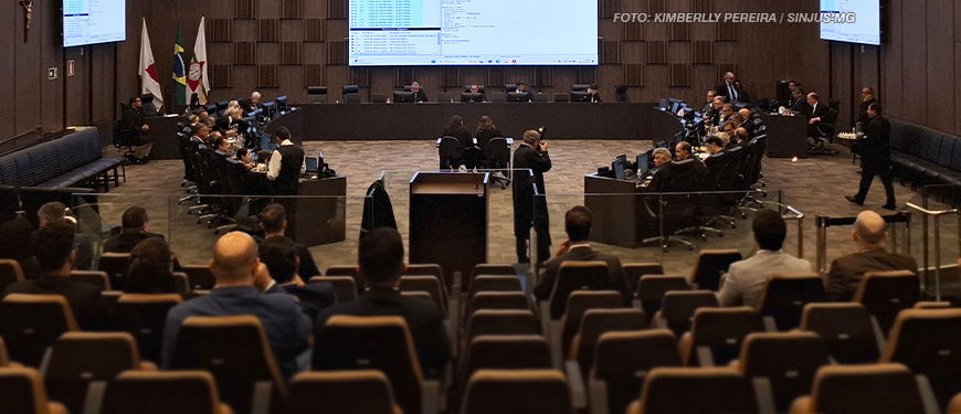 A imagem retrata um auditório formal, amplo e moderno, configurado para uma sessão oficial. Ao fundo, há um painel eletrônico com informações projetadas, acompanhado de uma bancada elevada onde estão reunidos membros do Órgão Especial. Nas laterais e no centro da sala, pessoas estão sentadas em mesas ou cadeiras, em atividades como anotações ou observação, enquanto outros estão em pé registrando o evento. Algumas bandeiras do Brasil e de estados estão dispostas à esquerda, reforçando o caráter institucional. A iluminação é clara, e o espaço está bem organizado, com cadeiras ocupadas por espectadores. A cena transmite o ambiente de uma deliberação oficial importante, destacando a aprovação de mudanças na lei do Auxílio-Saúde para a ALMG.