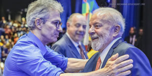 A imagem destaca o presidente Lula e o governador de Minas Gerais, Romeu Zema, cumprimentando-se de maneira cordial. Ambos parecem sorridentes e próximos, em um momento de cumprimento com um aperto de mãos. Ao fundo, vê-se o ambiente decorado e outras pessoas, com um clima que remete a um encontro público. As manchetes relacionadas fazem referência à adesão de Minas Gerais ao RRF e aos desdobramentos políticos envolvendo sindicatos que reivindicam a sanção do Propag e articulam estratégias de negociação