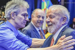 A imagem destaca o presidente Lula e o governador de Minas Gerais, Romeu Zema, cumprimentando-se de maneira cordial. Ambos parecem sorridentes e próximos, em um momento de cumprimento com um aperto de mãos. Ao fundo, vê-se o ambiente decorado e outras pessoas, com um clima que remete a um encontro público. As manchetes relacionadas fazem referência à adesão de Minas Gerais ao RRF e aos desdobramentos políticos envolvendo sindicatos que reivindicam a sanção do Propag e articulam estratégias de negociação