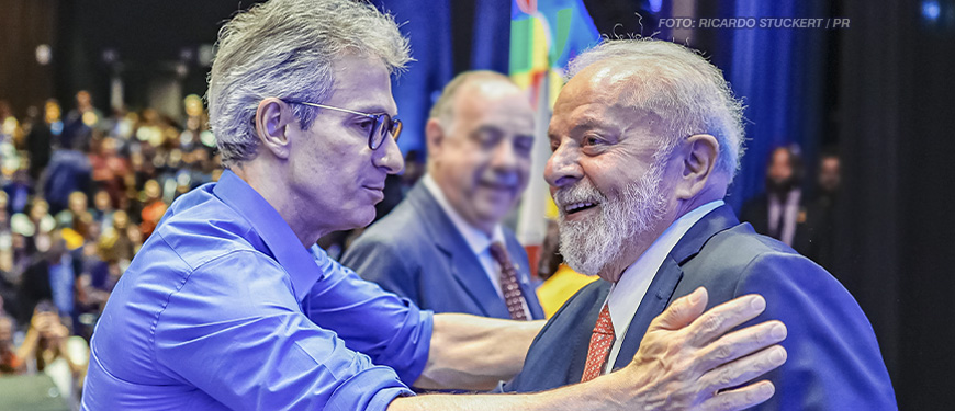 A imagem destaca o presidente Lula e o governador de Minas Gerais, Romeu Zema, cumprimentando-se de maneira cordial. Ambos parecem sorridentes e próximos, em um momento de cumprimento com um aperto de mãos. Ao fundo, vê-se o ambiente decorado e outras pessoas, com um clima que remete a um encontro público. As manchetes relacionadas fazem referência à adesão de Minas Gerais ao RRF e aos desdobramentos políticos envolvendo sindicatos que reivindicam a sanção do Propag e articulam estratégias de negociação