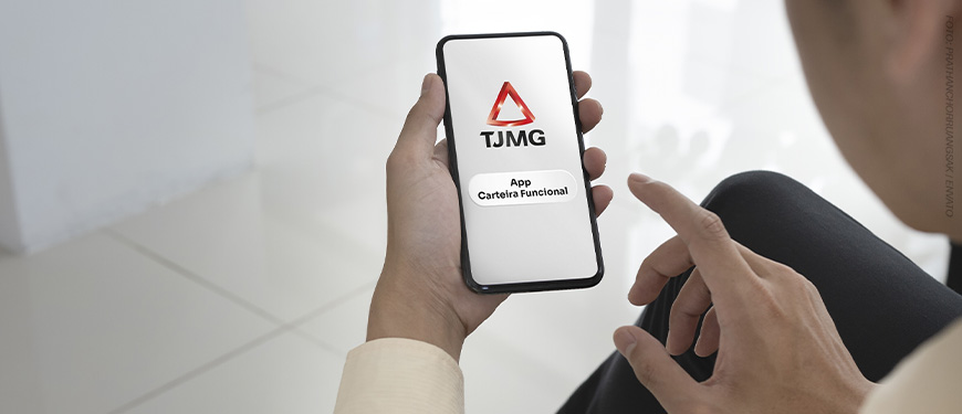 A imagem mostra uma pessoa segurando um smartphone, onde aparece a tela inicial do aplicativo da carteira funcional digital do Tribunal de Justiça de Minas Gerais (TJMG). O logo do TJMG está destacado na tela do celular. A cena tem um tom profissional e minimalista, com o fundo desfocado, dando ênfase ao telefone.