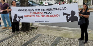 Em primeiro plano, há duas mulheres segurando uma faixa onde está escrito: "Aniversário sem comemoração, lutamos pelo direito da nomeação", Ao fundo estão árvores e uma vista da avenida onde fica a sede do TJMG. Conteúdo textual: Pauta prioritária - Aprovados do concurso 01/2022 do TJMG fazem ato por nomeações