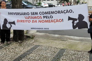 Em primeiro plano, há duas mulheres segurando uma faixa onde está escrito: "Aniversário sem comemoração, lutamos pelo direito da nomeação", Ao fundo estão árvores e uma vista da avenida onde fica a sede do TJMG. Conteúdo textual: Pauta prioritária - Aprovados do concurso 01/2022 do TJMG fazem ato por nomeações