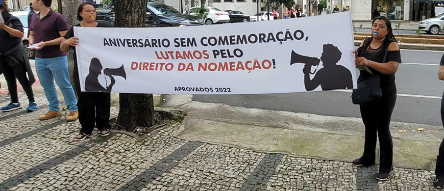 Em primeiro plano, há duas mulheres segurando uma faixa onde está escrito: "Aniversário sem comemoração, lutamos pelo direito da nomeação", Ao fundo estão árvores e uma vista da avenida onde fica a sede do TJMG. Conteúdo textual: Pauta prioritária - Aprovados do concurso 01/2022 do TJMG fazem ato por nomeações