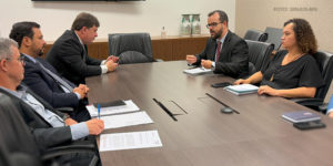 A imagem mostra uma reunião em uma sala de conferências com uma mesa grande de madeira escura, com documentos e canetas sobre ela, cercada por cadeiras pretas. Representantes do SINJUS-MG, do SERJUSMIG e do SINDOJUS e da Administração do TJMG participam ativamente da conversa, com cadernos e anotações à mão, discutindo problemas relacionados ao DJEN. O ambiente é formal e bem iluminado, com uma televisão desligada na parede e alguns objetos decorativos no fundo.