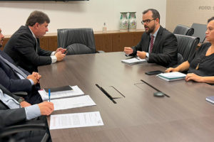 A imagem mostra uma reunião em uma sala de conferências com uma mesa grande de madeira escura, com documentos e canetas sobre ela, cercada por cadeiras pretas. Representantes do SINJUS-MG, do SERJUSMIG e do SINDOJUS e da Administração do TJMG participam ativamente da conversa, com cadernos e anotações à mão, discutindo problemas relacionados ao DJEN. O ambiente é formal e bem iluminado, com uma televisão desligada na parede e alguns objetos decorativos no fundo.