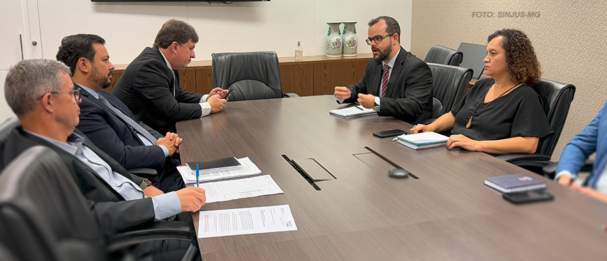 A imagem mostra uma reunião em uma sala de conferências com uma mesa grande de madeira escura, com documentos e canetas sobre ela, cercada por cadeiras pretas. Representantes do SINJUS-MG, do SERJUSMIG e do SINDOJUS e da Administração do TJMG participam ativamente da conversa, com cadernos e anotações à mão, discutindo problemas relacionados ao DJEN. O ambiente é formal e bem iluminado, com uma televisão desligada na parede e alguns objetos decorativos no fundo.