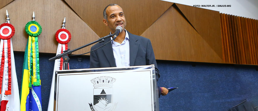 Nesta imagem, está Wagner Ferreira, um homem negro de cabelos grisalhos bem curtos, ele está em pé atrás de um púlpito, vestindo um paletó preto e camisa clara, falando ao microfone. Ao fundo, vemos três bandeiras, a do Brasil, a do estado de Minas Gerais e de Belo Horizonte. O brasão da cidade está visível na frente do púlpito, identificando a Câmara Municipal.