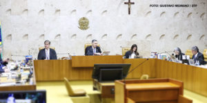 As imagens mostram uma sessão no plenário do STF do Brasil, com ministros sentados em uma bancada semicircular posicionados em cadeiras de madeira. Ao fundo, na parede, está o brasão do Brasil e uma cruz. Alguns monitores exibem uma pessoa discursando, e há pessoas na plateia assistindo à sessão. Em duas das imagens, aparece sobreposto um texto em letras grandes, destacando a expressão "Nota pública à decisão do STF sobre o regime jurídico único: um erro fatal" em vermelho e preto, enfatizando uma crítica sobre a decisão do tribunal.