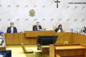 As imagens mostram uma sessão no plenário do STF do Brasil, com ministros sentados em uma bancada semicircular posicionados em cadeiras de madeira. Ao fundo, na parede, está o brasão do Brasil e uma cruz. Alguns monitores exibem uma pessoa discursando, e há pessoas na plateia assistindo à sessão. Em duas das imagens, aparece sobreposto um texto em letras grandes, destacando a expressão "Nota pública à decisão do STF sobre o regime jurídico único: um erro fatal" em vermelho e preto, enfatizando uma crítica sobre a decisão do tribunal.