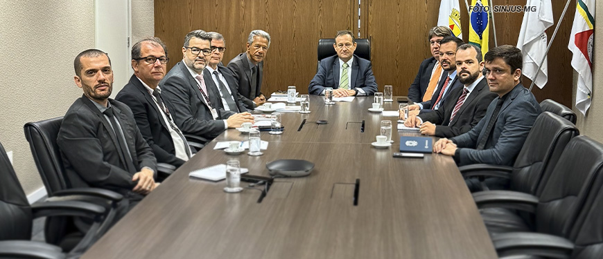 As imagens mostram uma reunião formal com onze pessoas sentadas ao redor de uma mesa retangular em uma sala de reuniões. Os participantes, todos homens, estão vestidos de terno e gravata, com expressões sérias e postura formal. Ao fundo, há uma parede de madeira com bandeiras do Brasil e de outras instituições à direita. A legenda indica que o Tribunal de Justiça enviará uma proposta de Data-Base 2024 para a ALMG nos próximos dias, além de outras definições. O ambiente transmite um clima profissional e institucional.