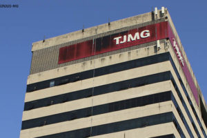 Fotografia diurna da fachada do TJMG, um prédio na cor bege com janelas horizontais, onde se vê acima delas uma placa vermelha com letras brancas (TJMG).