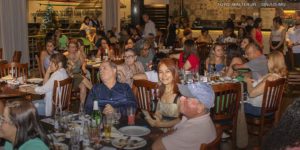 As imagens mostram um evento de confraternização em um restaurante, com diversas pessoas sentadas em mesas, conversando e aproveitando um momento de integração. As mesas estão repletas de pratos e copos. O ambiente é aconchegante, com iluminação interna e uma decoração acolhedora. Em uma das fotos, algumas pessoas estão em pé diante de um banner decorativo, realizando um discurso ou apresentação. A atmosfera geral sugere um encontro descontraído e amigável, reforçando os laços entre os servidores do NAP e NPD.