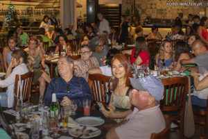 As imagens mostram um evento de confraternização em um restaurante, com diversas pessoas sentadas em mesas, conversando e aproveitando um momento de integração. As mesas estão repletas de pratos e copos. O ambiente é aconchegante, com iluminação interna e uma decoração acolhedora. Em uma das fotos, algumas pessoas estão em pé diante de um banner decorativo, realizando um discurso ou apresentação. A atmosfera geral sugere um encontro descontraído e amigável, reforçando os laços entre os servidores do NAP e NPD.