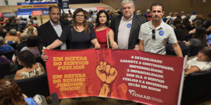 A imagem mostra um grupo de cinco pessoas segurando uma faixa vermelha em um auditório lotado, durante o seminário na Câmara dos Deputados. A faixa tem mensagens de apoio ao serviço público e ao Regime Jurídico Único, destacando a importância da estabilidade para garantir continuidade, imparcialidade e impessoalidade no serviço público. O evento discute o possível fim desse regime, e várias pessoas no auditório estão acompanhando a discussão. A Fenajud, entidade que representa servidores do Judiciário, está envolvida na mobilização.