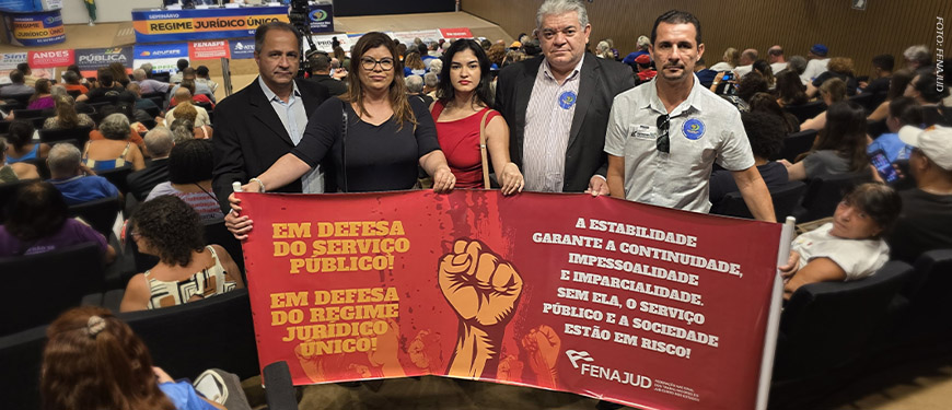 A imagem mostra um grupo de cinco pessoas segurando uma faixa vermelha em um auditório lotado, durante o seminário na Câmara dos Deputados. A faixa tem mensagens de apoio ao serviço público e ao Regime Jurídico Único, destacando a importância da estabilidade para garantir continuidade, imparcialidade e impessoalidade no serviço público. O evento discute o possível fim desse regime, e várias pessoas no auditório estão acompanhando a discussão. A Fenajud, entidade que representa servidores do Judiciário, está envolvida na mobilização.