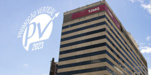 Fotografia de um grande prédio com fachada bege e janelas espelhadas escuras. No topo do edifício, há placas vermelhas com a sigla "TJMG" (Tribunal de Justiça de Minas Gerais). O céu azul com algumas nuvens aparece ao fundo. Na parte superior da imagem, há um logotipo branco com a inscrição "Promoção Vertical PV 2023" e uma seta apontando para cima. Conteúdo textual: CARREIRA - TJMG divulga decisão acerca dos pedidos de reconsideração da PV 2023.