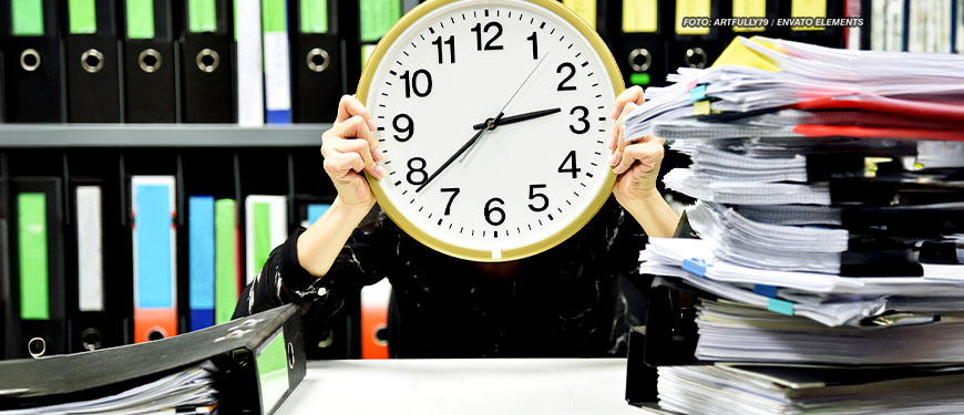 Uma pessoa vestida com camisa preta está segurando um relógio grande na frente de seu rosto, com prateleiras de arquivos e uma pilha de papéis ao fundo.