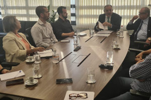 A imagem mostra uma reunião com várias pessoas sentadas ao redor de uma mesa, discutindo, estão presentes na reunião o vice-presidente do TJMG, Marcos Lincoln dos Santos com sua equipe e os dirigentes do SINJUS-MG, Alexandre Pires (coordenador-geral) e Felipe Rodrigues (diretor de Assuntos Jurídicos). No canto inferior esquerdo, há um selo com a hashtag "#RevogaA17".