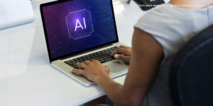 Uma pessoa está sentada à mesa, trabalhando em um laptop. Na tela do laptop, aparecem as letras "AI" (Inteligência Artificial) no centro, rodeadas por um design digital e futurista, lembrando circuitos eletrônicos.