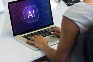 Uma pessoa está sentada à mesa, trabalhando em um laptop. Na tela do laptop, aparecem as letras "AI" (Inteligência Artificial) no centro, rodeadas por um design digital e futurista, lembrando circuitos eletrônicos.