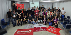 Grupo grande de pessoas reunidas em uma sala de conferências com cadeiras organizadas ao redor. Há uma tela de projeção ao fundo e várias bandeiras de sindicatos no chão, incluindo as do SERJUSMIG, SINJUS-MG, FENAJUD, SINDIJUS, entre outras. Estão no grupo o coordenador-geral do SINJUS-MG, Alexandre Pires, e o diretor de Assuntos Jurídicos, Felipe Rodrigues. É visível um ambiente de diversidade, com pessoas de diferentes idades, gêneros e estilos, indicando um encontro de representatividade sindical e compromisso com uma causa coletiva.