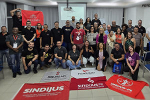 Grupo grande de pessoas reunidas em uma sala de conferências com cadeiras organizadas ao redor. Há uma tela de projeção ao fundo e várias bandeiras de sindicatos no chão, incluindo as do SERJUSMIG, SINJUS-MG, FENAJUD, SINDIJUS, entre outras. Estão no grupo o coordenador-geral do SINJUS-MG, Alexandre Pires, e o diretor de Assuntos Jurídicos, Felipe Rodrigues. É visível um ambiente de diversidade, com pessoas de diferentes idades, gêneros e estilos, indicando um encontro de representatividade sindical e compromisso com uma causa coletiva.