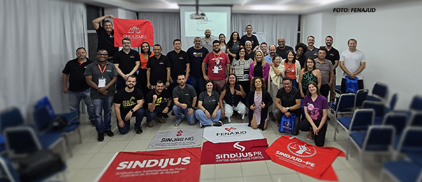 Grupo grande de pessoas reunidas em uma sala de conferências com cadeiras organizadas ao redor. Há uma tela de projeção ao fundo e várias bandeiras de sindicatos no chão, incluindo as do SERJUSMIG, SINJUS-MG, FENAJUD, SINDIJUS, entre outras. Estão no grupo o coordenador-geral do SINJUS-MG, Alexandre Pires, e o diretor de Assuntos Jurídicos, Felipe Rodrigues. É visível um ambiente de diversidade, com pessoas de diferentes idades, gêneros e estilos, indicando um encontro de representatividade sindical e compromisso com uma causa coletiva.