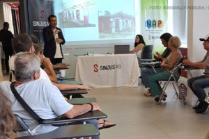 A imagem captura um momento de reunião do NAP em uma sala iluminada. Em pé na frente da sala está Marcos Haddad de terno escuro, falando para o grupo de participantes sentados em cadeiras organizadas em semicírculo. Ao fundo, uma tela de projeção exibe imagens, e banners do SINJUS-MG e do NAP estão posicionados ao lado de uma mesa coberta com materiais. Os participantes escutam atentamente enquanto seguram copos e garrafas de água. O texto na imagem destaca que a reunião apresenta o planejamento de eventos e viagens para 2025.