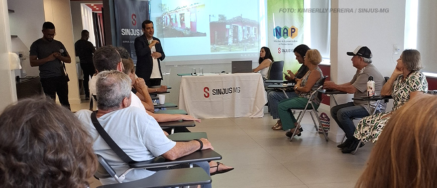 A imagem captura um momento de reunião do NAP em uma sala iluminada. Em pé na frente da sala está Marcos Haddad de terno escuro, falando para o grupo de participantes sentados em cadeiras organizadas em semicírculo. Ao fundo, uma tela de projeção exibe imagens, e banners do SINJUS-MG e do NAP estão posicionados ao lado de uma mesa coberta com materiais. Os participantes escutam atentamente enquanto seguram copos e garrafas de água. O texto na imagem destaca que a reunião apresenta o planejamento de eventos e viagens para 2025.