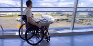 Um homem em uma cadeira de rodas está em um aeroporto, observando através de uma grande parede de vidro um avião estacionado na pista. Ele veste uma camisa clara e calça bege. O céu está azul com algumas nuvens, e a estrutura do aeroporto é moderna, com amplas janelas que permitem a visão do lado externo, onde há mais aviões e equipamentos de solo.