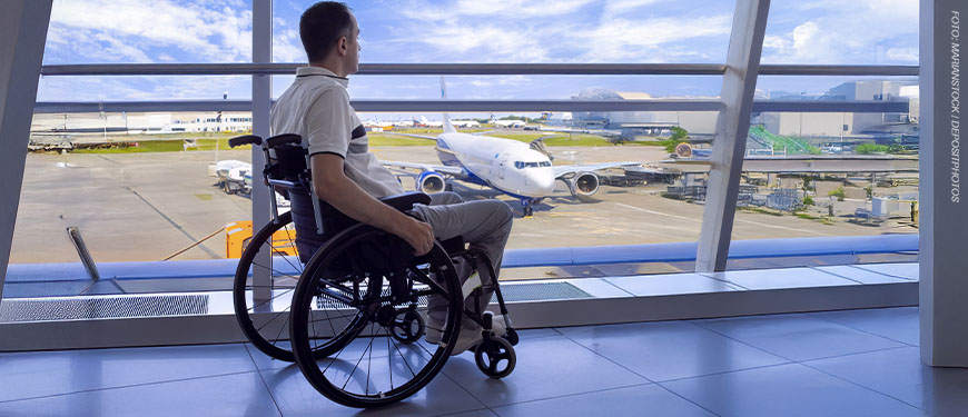 Um homem em uma cadeira de rodas está em um aeroporto, observando através de uma grande parede de vidro um avião estacionado na pista. Ele veste uma camisa clara e calça bege. O céu está azul com algumas nuvens, e a estrutura do aeroporto é moderna, com amplas janelas que permitem a visão do lado externo, onde há mais aviões e equipamentos de solo.