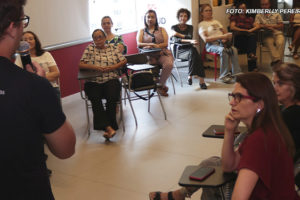 A imagem mostra um grupo de pessoas em uma sala de aula ou reunião, onde o NAP discute os benefícios da atividade física para pessoas com mais de 50 anos. Vários participantes estão sentados, atentos, enquanto um palestrante com microfone apresenta o conteúdo, todos engajados e interessados no tema. Em uma imagem adicional, vemos um grupo de pessoas, incluindo instrutores e participantes, posando sorridentes.