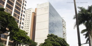 A imagem exibe a fachada do prédio localizado na Avenida Afonso Pena 1.500 em Belo Horizonte. O edifício possui uma lateral de concreto desgastado e outra com grandes janelas espelhadas. Em primeiro plano, há uma avenida movimentada com carros e árvores alinhadas ao longo da calçada. O céu está nublado, e o tom da imagem transmite um ar de seriedade. O texto na imagem informa que a atuação do SINJUS garantiu uma resposta do TJMG sobre a insalubridade no edifício