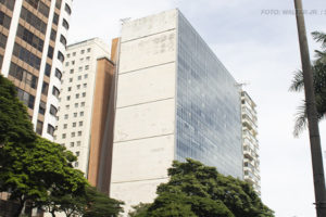 A imagem exibe a fachada do prédio localizado na Avenida Afonso Pena 1.500 em Belo Horizonte. O edifício possui uma lateral de concreto desgastado e outra com grandes janelas espelhadas. Em primeiro plano, há uma avenida movimentada com carros e árvores alinhadas ao longo da calçada. O céu está nublado, e o tom da imagem transmite um ar de seriedade. O texto na imagem informa que a atuação do SINJUS garantiu uma resposta do TJMG sobre a insalubridade no edifício