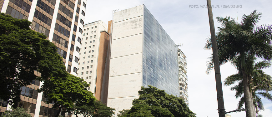 A imagem exibe a fachada do prédio localizado na Avenida Afonso Pena 1.500 em Belo Horizonte. O edifício possui uma lateral de concreto desgastado e outra com grandes janelas espelhadas. Em primeiro plano, há uma avenida movimentada com carros e árvores alinhadas ao longo da calçada. O céu está nublado, e o tom da imagem transmite um ar de seriedade. O texto na imagem informa que a atuação do SINJUS garantiu uma resposta do TJMG sobre a insalubridade no edifício